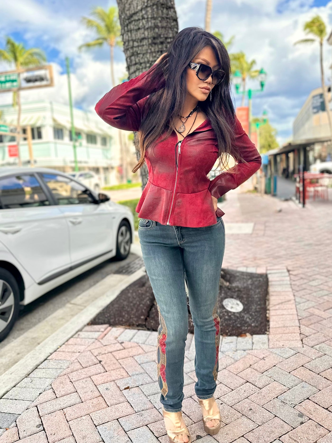 Woman in a red jacket and blue jeans standing on a sidewalk with palm trees in the background.