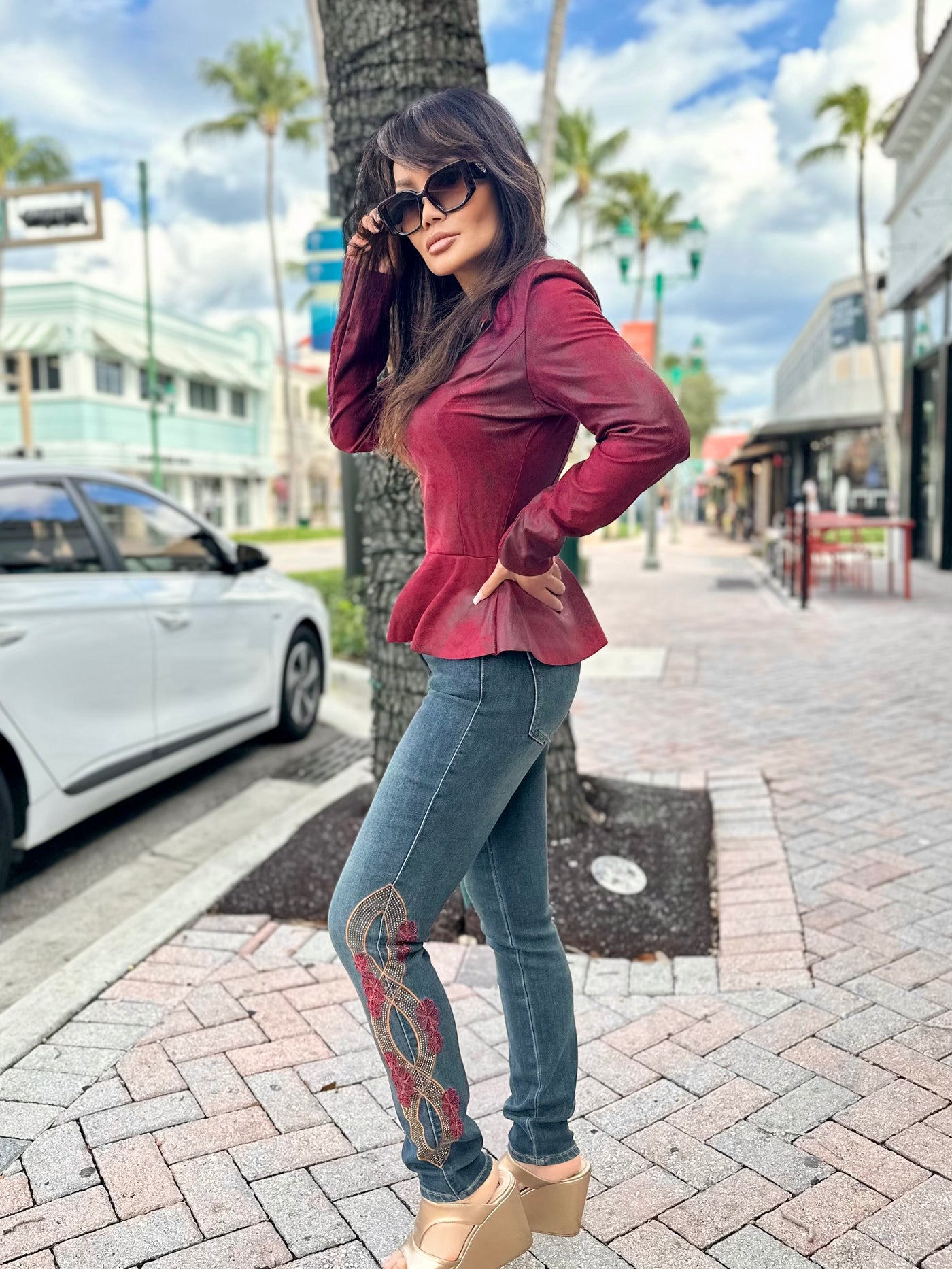 Woman in a red blouse and blue jeans standing on a sidewalk with palm trees in the background.