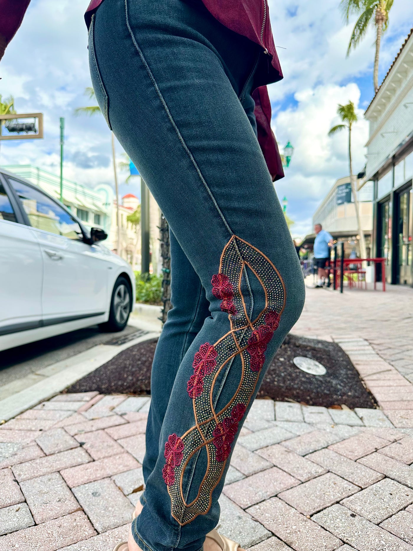 Person wearing blue jeans with red floral embroidery on a sidewalk.