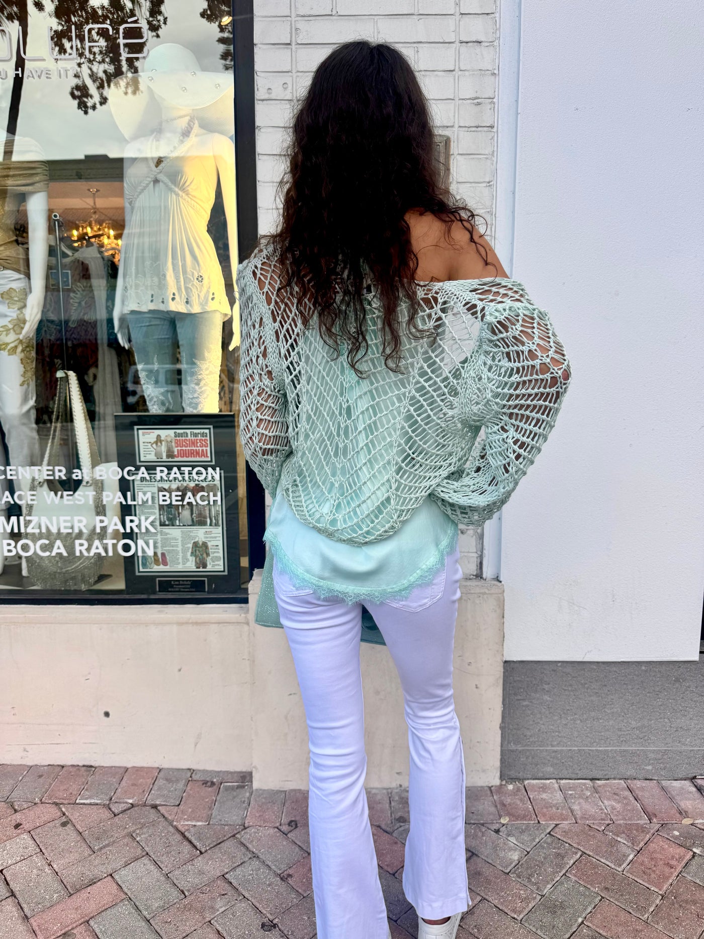 Woman wearing a light green crochet top and white pants standing in front of a store window display.