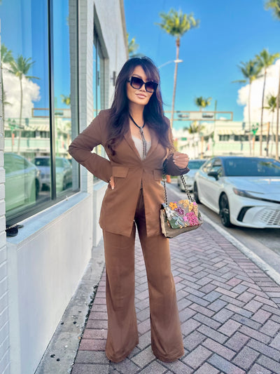 Woman in a brown suit standing on a sidewalk with palm trees and cars in the background