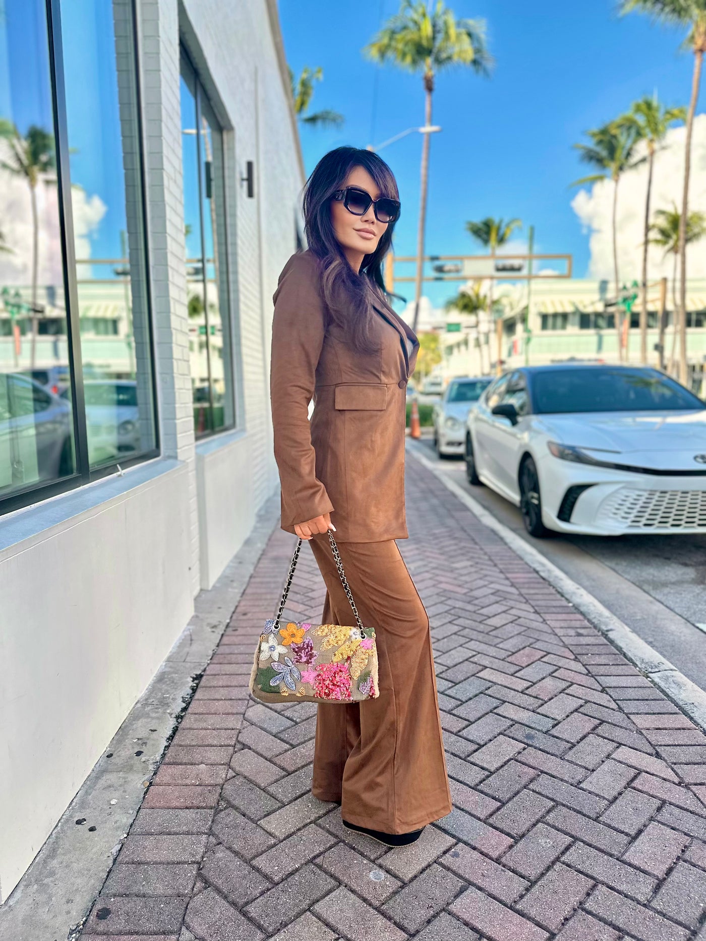 Woman in a brown suit holding a floral handbag on a sidewalk with palm trees in the background.