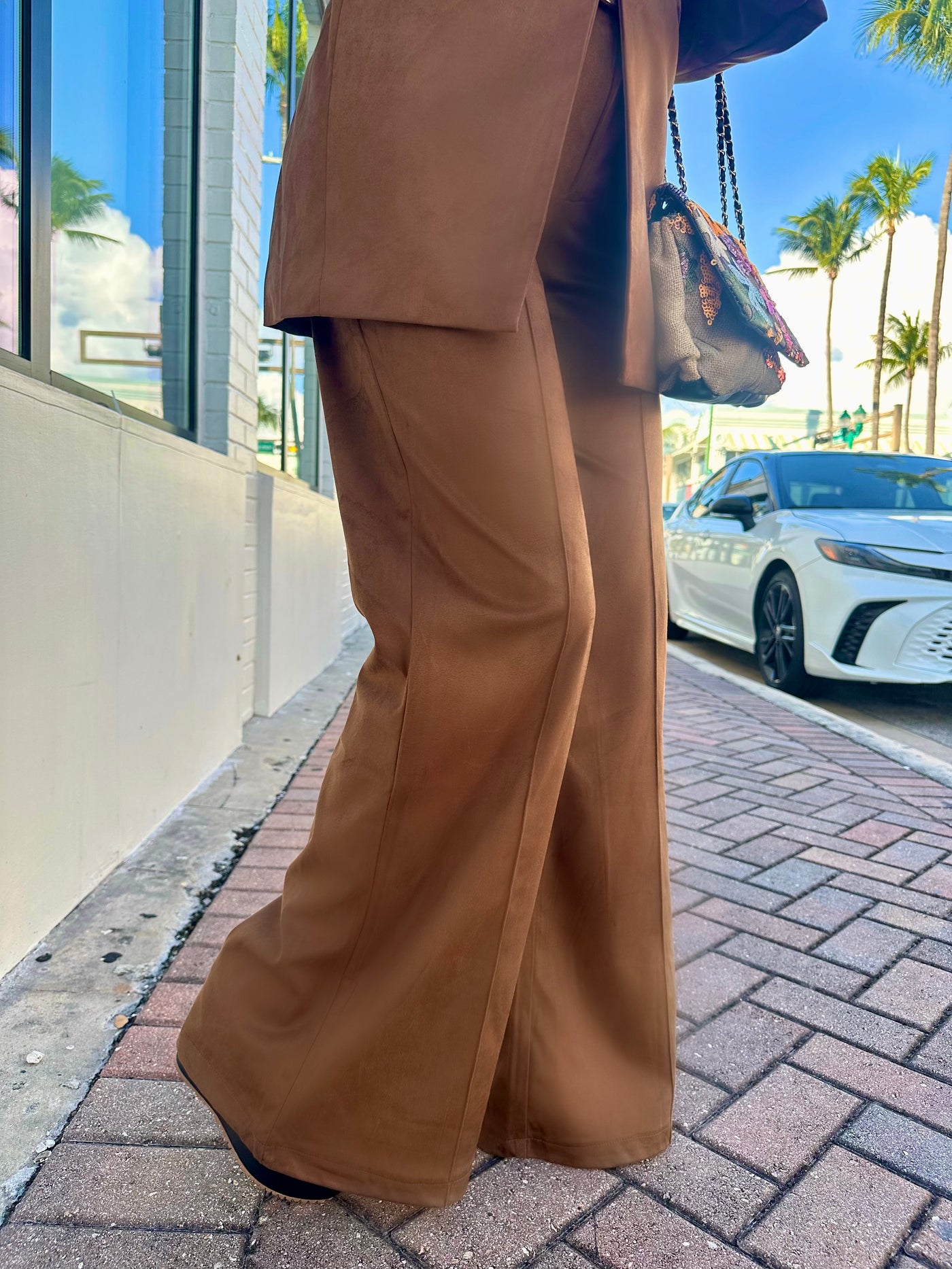 Person wearing a brown suit walking on a sidewalk with palm trees and a car in the background.