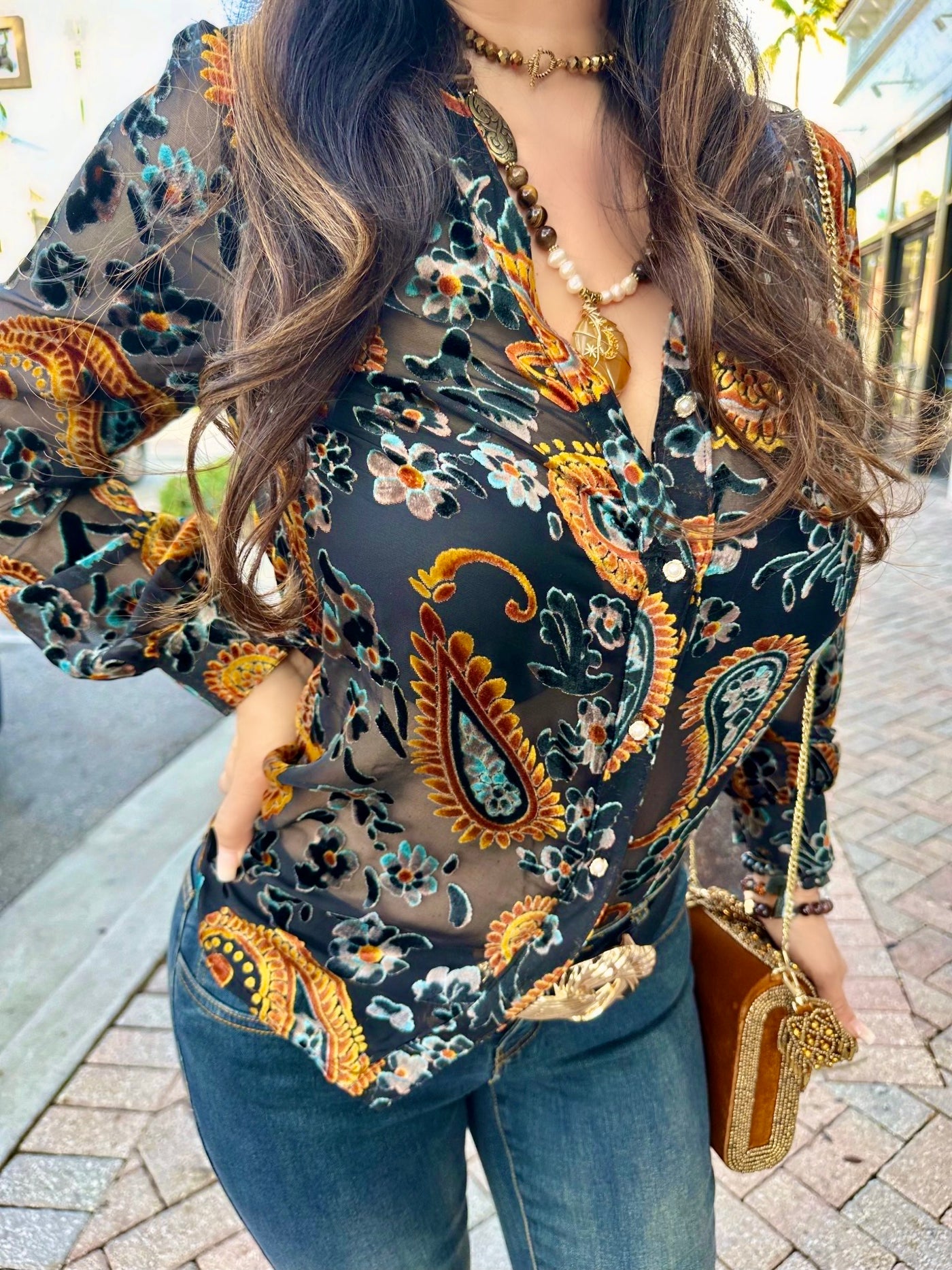 Woman wearing a patterned blouse with long sleeves and jeans, holding a gold clutch.
